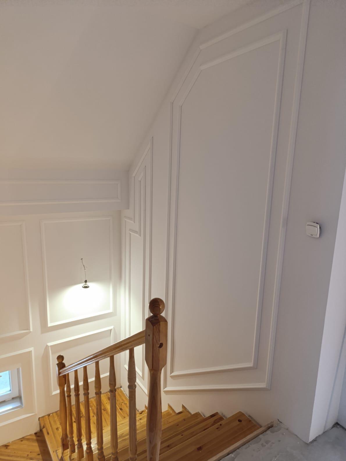 Klatka schodowa z drewnianymi schodami i białymi ścianami zdobionymi sztukaterią. Widoczne detale wykończenia, oświetlenie i fragment okna.