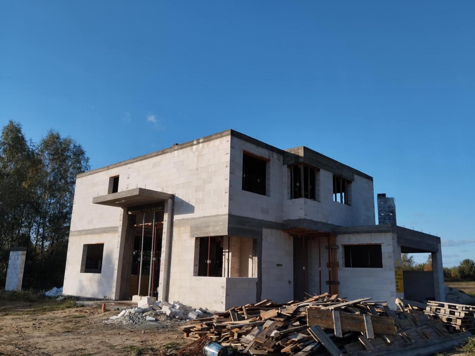 Surowy stan dwukondygnacyjnego domu z betonu komórkowego, widoczne otwory okienne i drzwiowe, podłoże z gruzem, błękitne niebo w tle.
