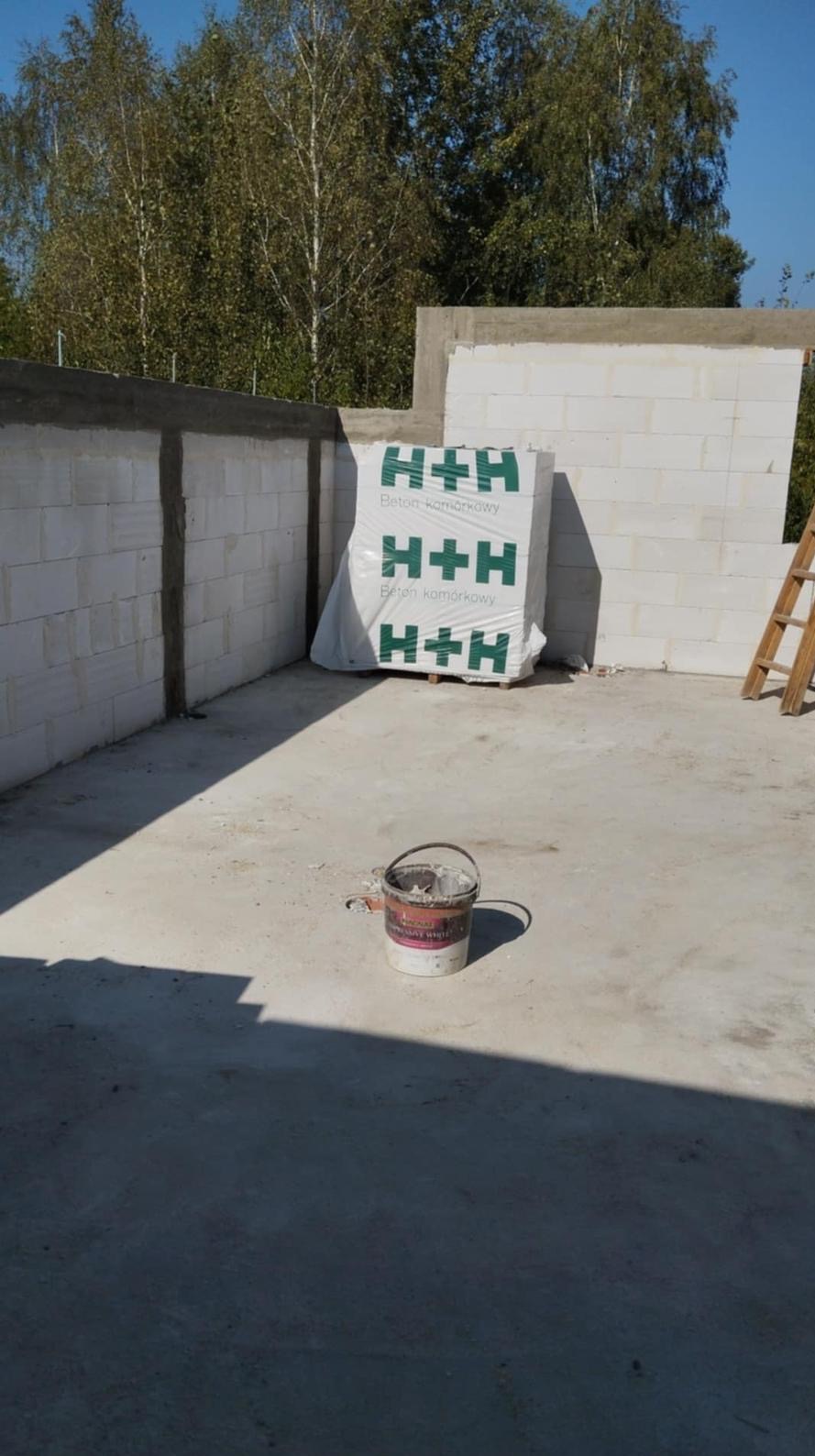 Budowa: Ściana z betonu komórkowego H+H w trakcie wznoszenia, widoczny kubeł z zaprawą i drabina oparta o ścianę. Tło: drzewa i błękitne niebo.