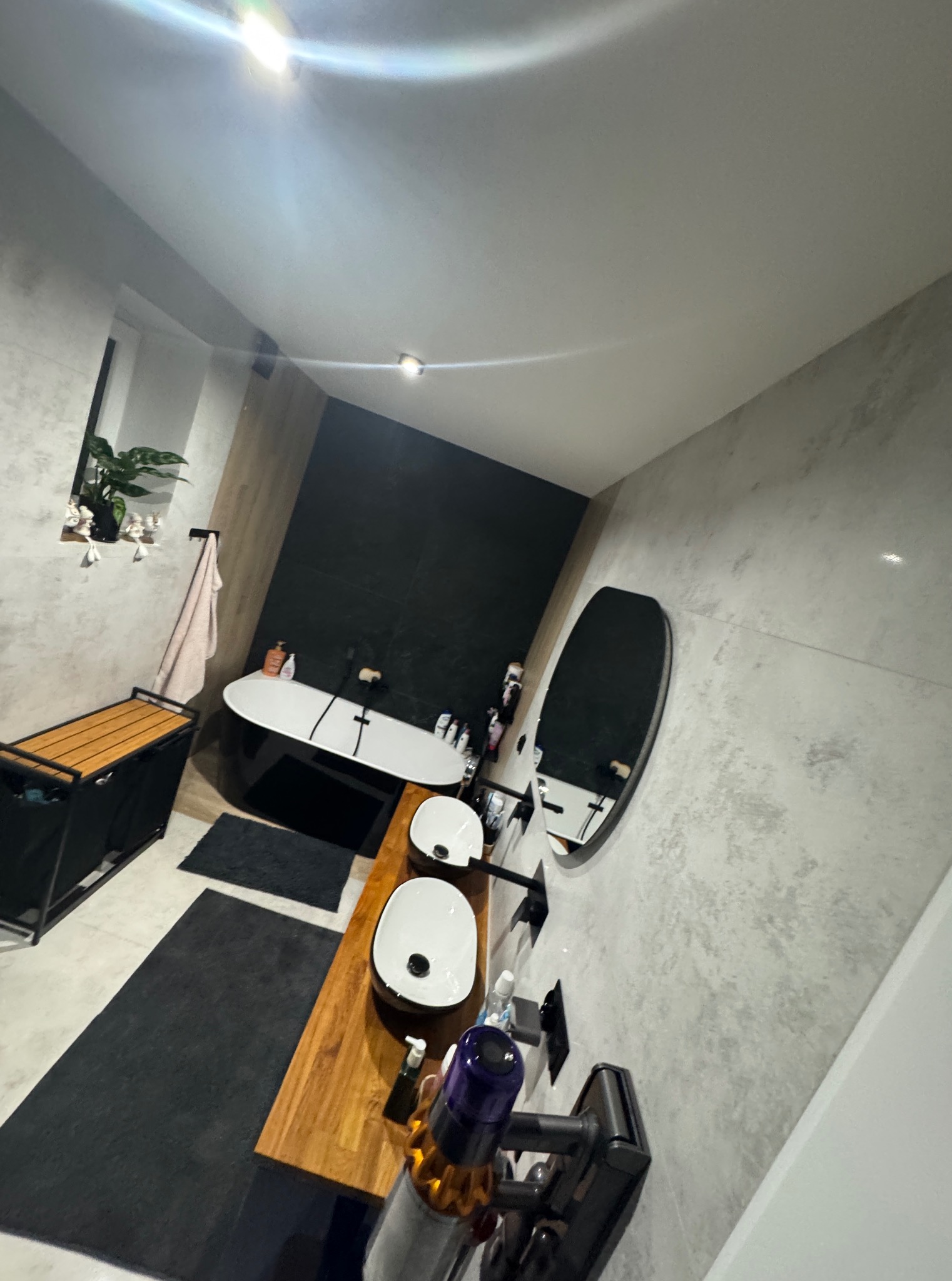 Nowoczesna łazienka z owalną wanną, drewnianym blatem pod umywalki i okrągłym lustrem. Szaro-białe płytki na ścianach, czarne akcenty i roślina w oknie.