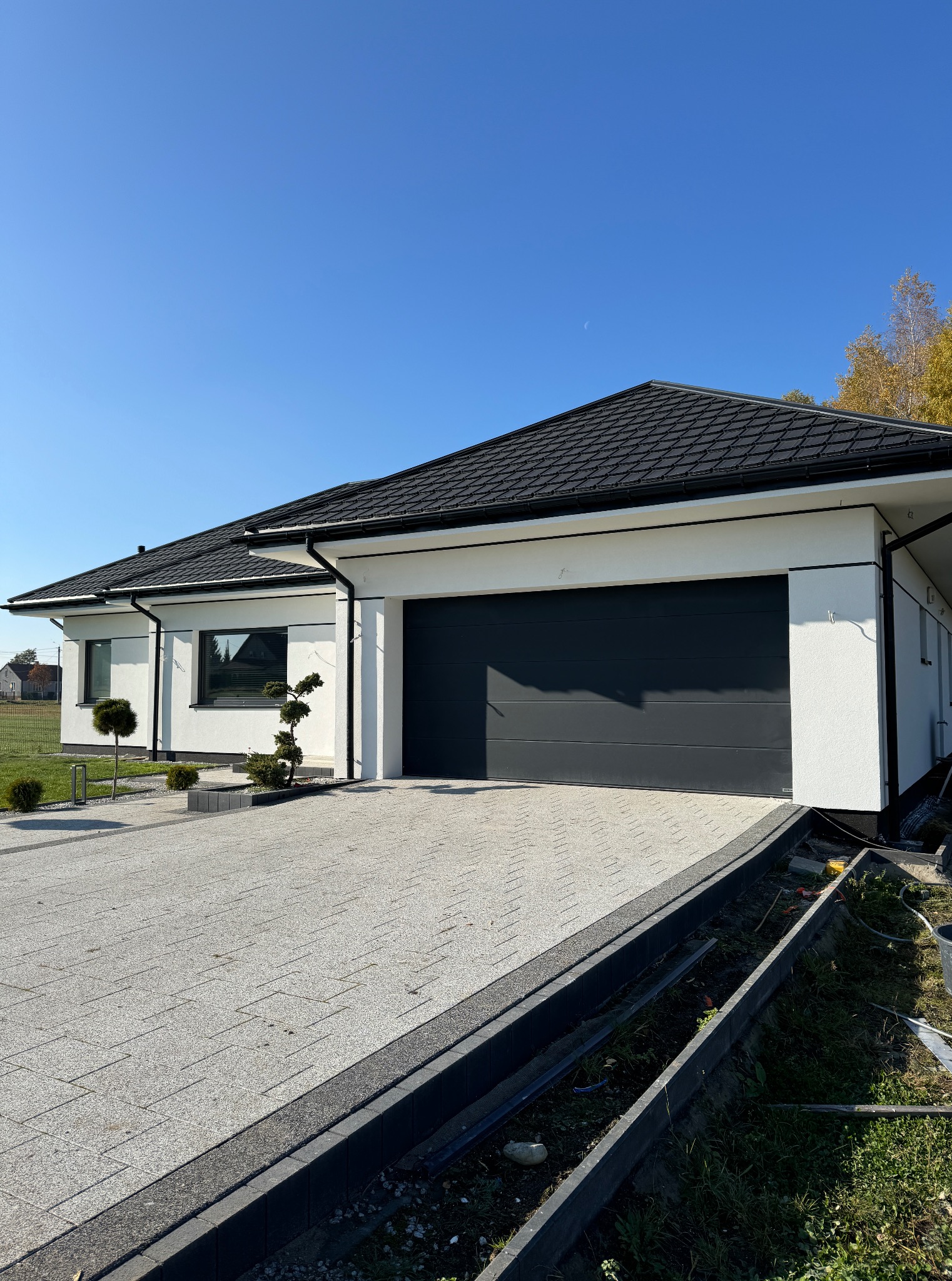 Nowoczesny dom jednorodzinny z grafitową bramą garażową i kostką brukową na podjeździe w słoneczny dzień. Widoczne detale wykończenia elewacji i zagospodarowania terenu.