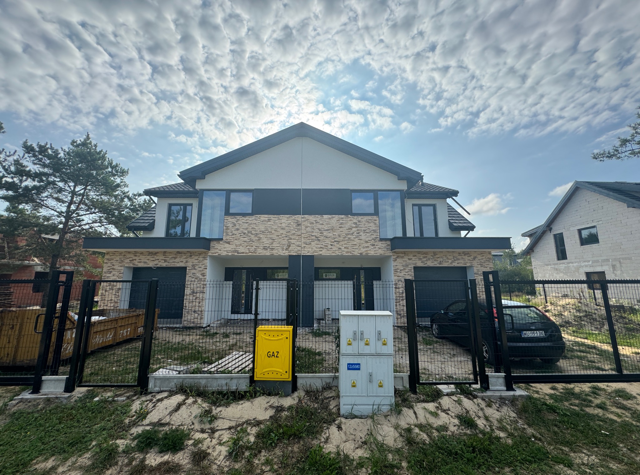 Dwukondygnacyjny bliźniak z jasną elewacją i elementami z imitacji cegły, widziany zza czarnego ogrodzenia. Na pierwszym planie skrzynka gazowa i elektryczna.