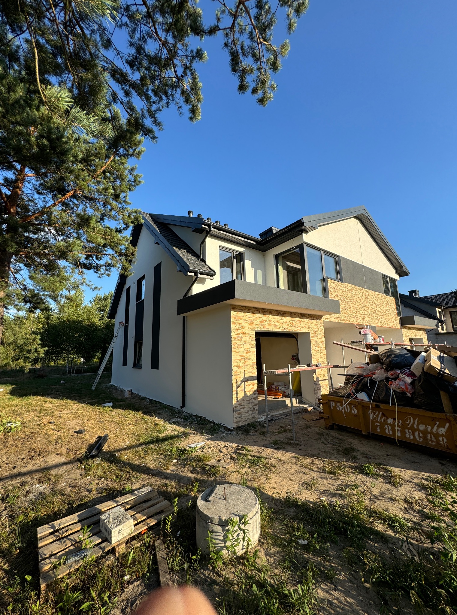 Dwukondygnacyjny dom z oknami w kolorze antracytu, wykończony beżowym tynkiem i kamieniem dekoracyjnym, w trakcie prac wykończeniowych. Na pierwszym planie kontener na odpady.