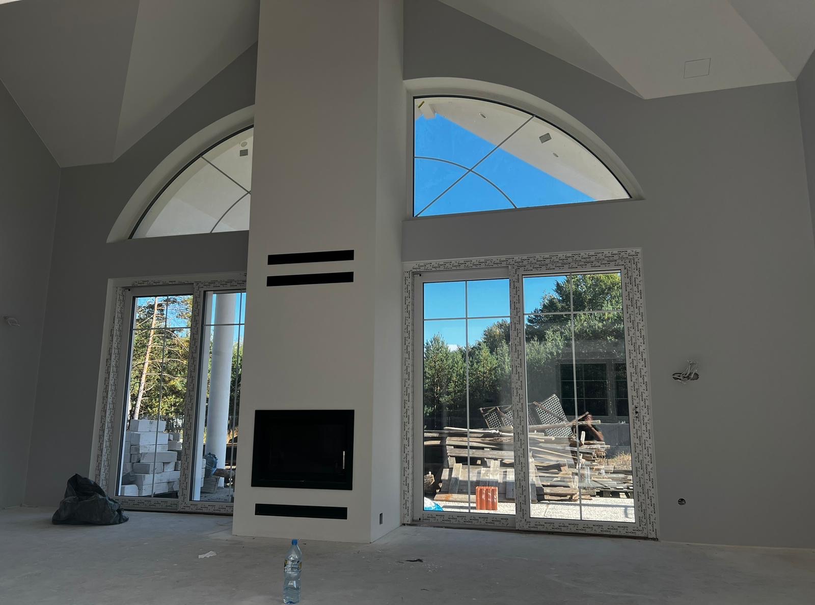 Wnętrze w trakcie wykańczania: szare ściany, duże okna z widokiem na zieleń, kominek z czarnym wkładem. Na podłodze butelka wody, za oknem materiały budowlane.
