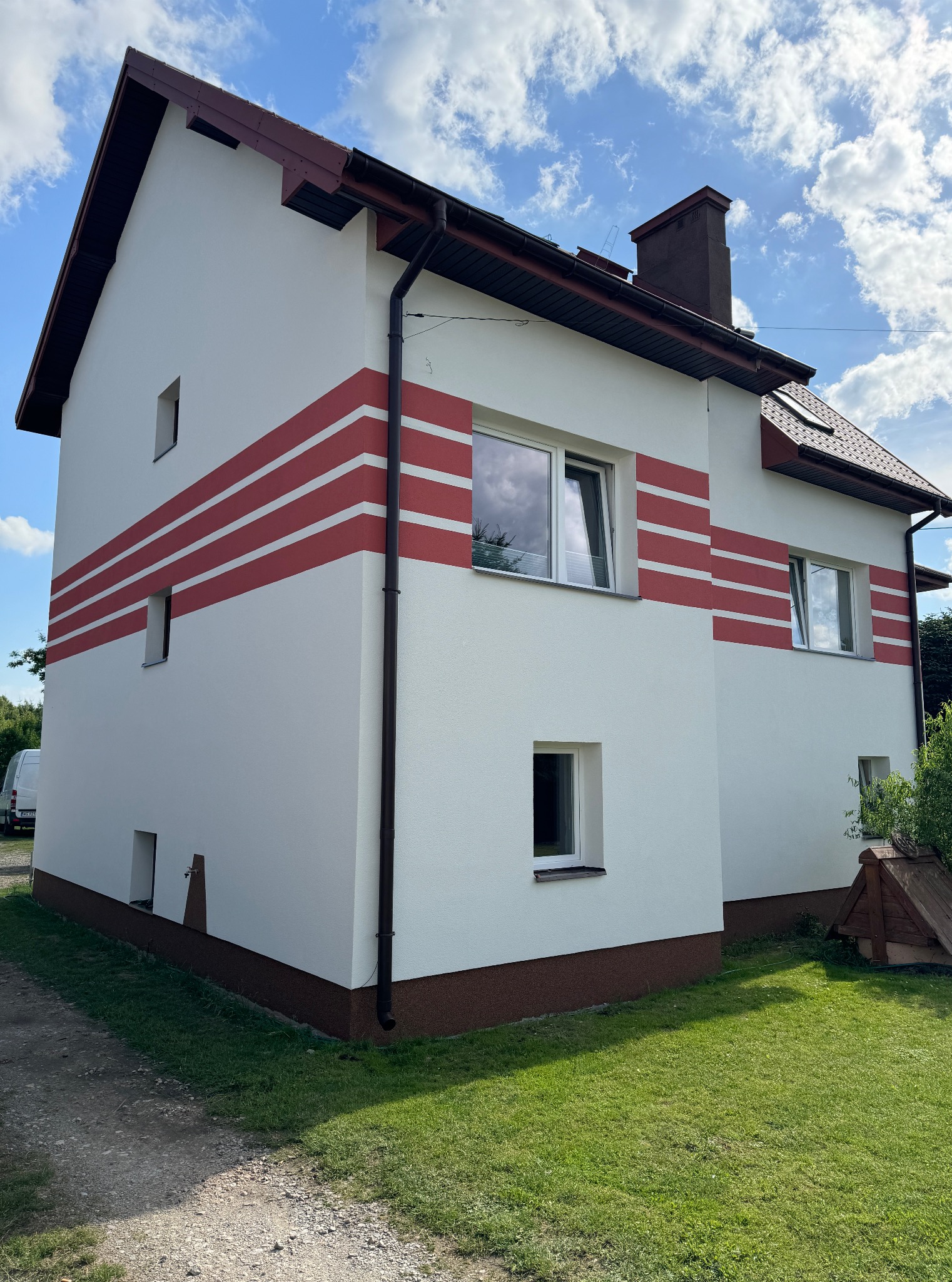 Dwupiętrowy dom z białą elewacją i czerwonymi pasami, brązowym dachem i rynnami. Widok na fragment ogrodu i niebo z chmurami.