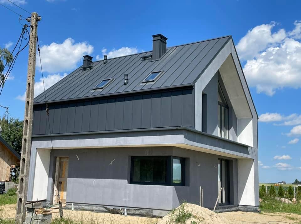 Nowoczesny dom z szarą blachą na dachu i oknami dachowymi, elewacja w trakcie wykończenia, widoczne elementy rusztowania i składowane materiały budowlane.