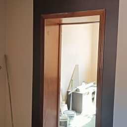 Arkadiusz Motyka - Dwukolorowe malowanie ścian z brązowym pasem obok drewnianej ościeżnicy. Widoczne narzędzia i pędzle malarskie. Jasne wnętrze w tle. Prace wykończeniowe w pomieszczeniu.