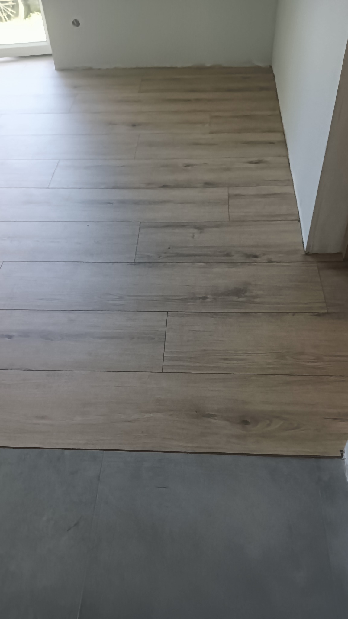 Ułożone panele podłogowe imitujące drewno w jasnym odcieniu, kontrastujące z szarą podłogą z płytek. Widok na fragment podłogi przy białej ścianie.