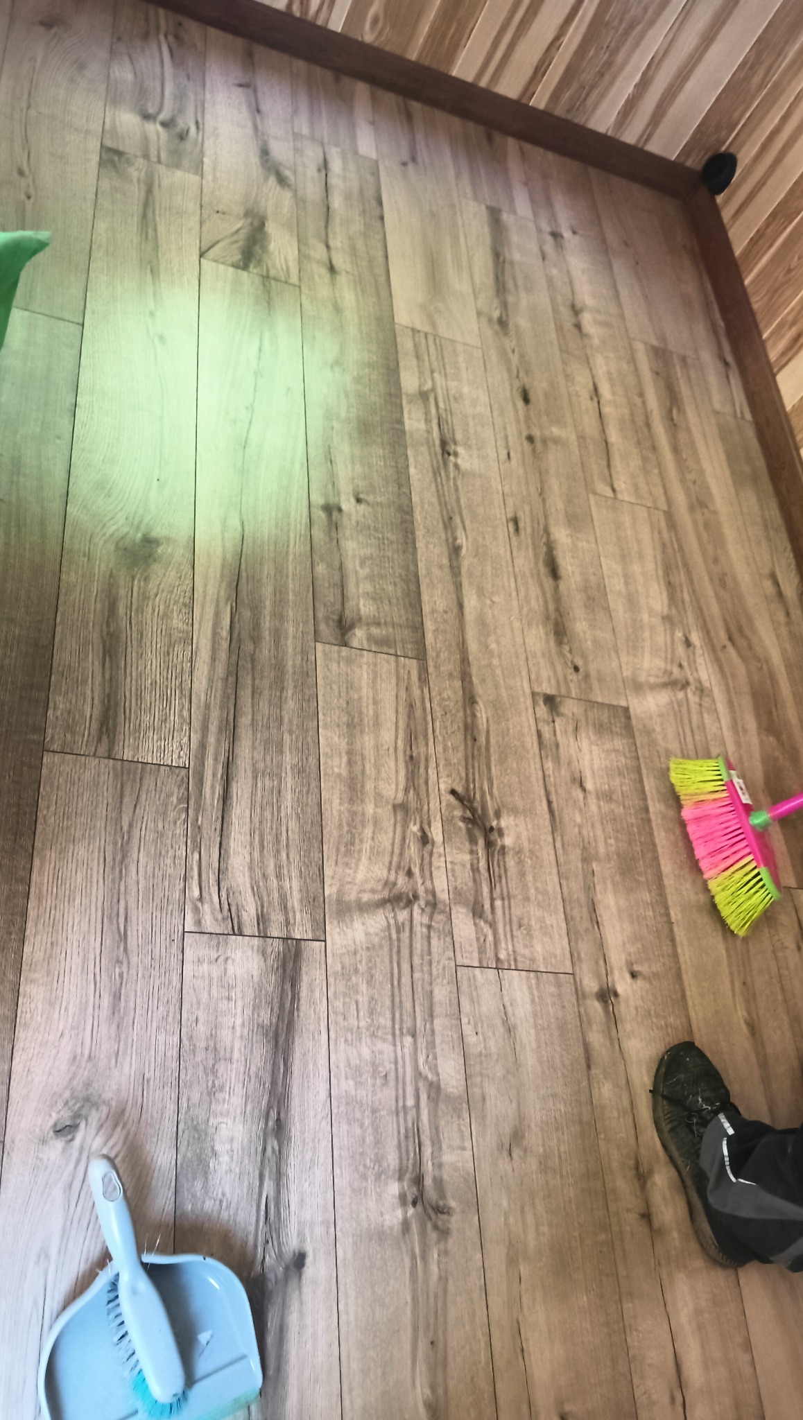 Parkiet z wzorem drewna, świeżo po montażu, z widocznymi narzędziami do sprzątania (szczotka, zmiotka). Ukończona podłoga w jasnych odcieniach brązu.