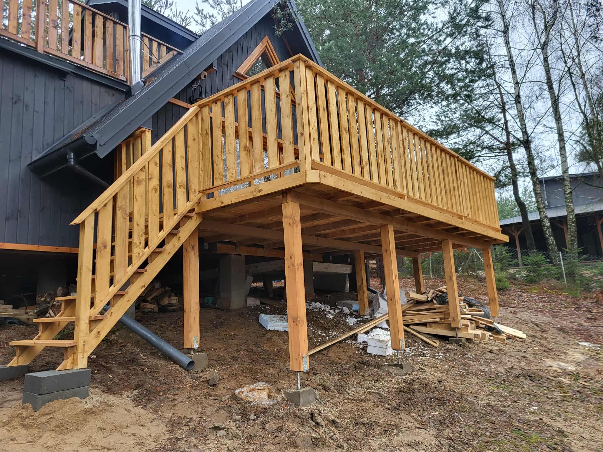 Drewniany taras z barierkami i schodami przy czarnym domu z kominem. Widoczne podpory tarasu na betonowych fundamentach i drewniane odpady budowlane.