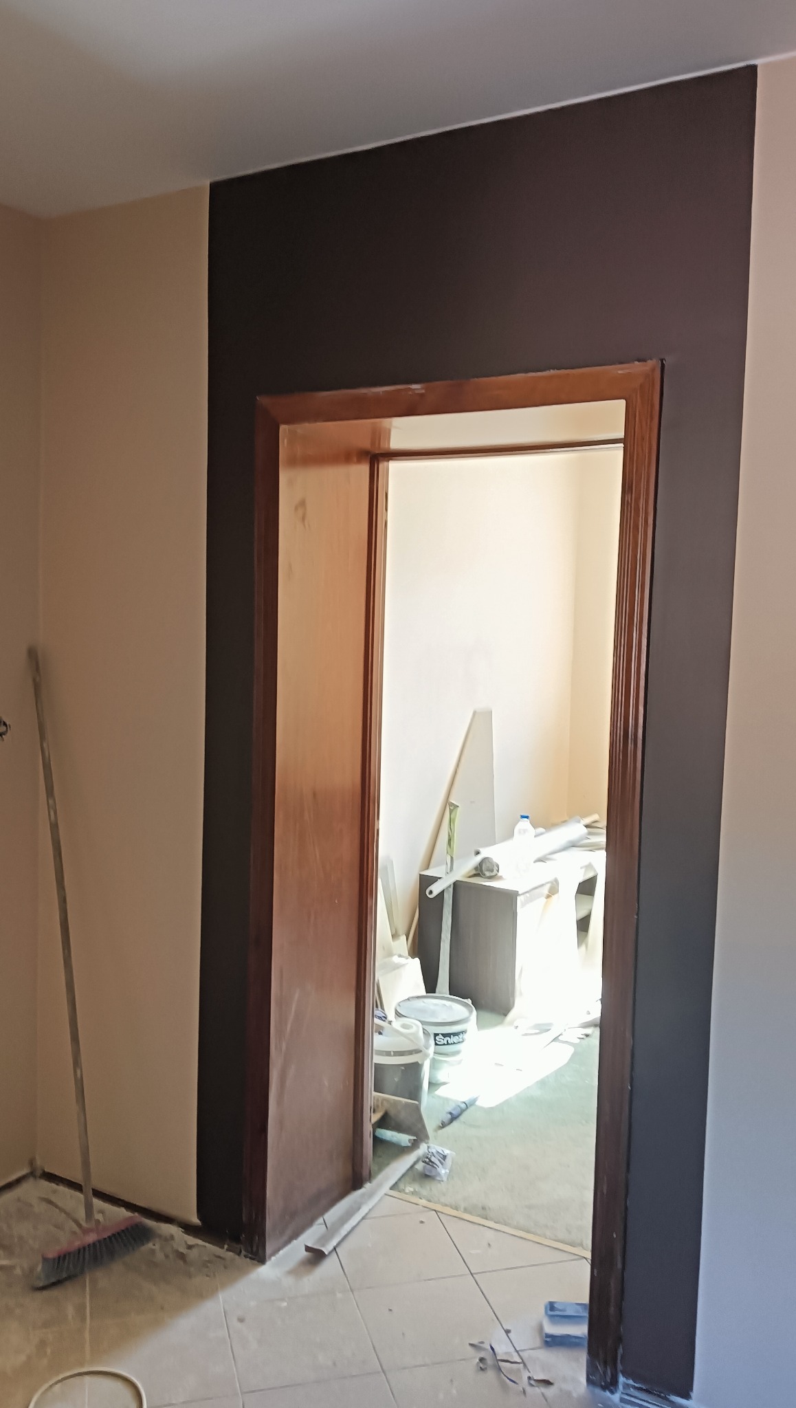 Dwukolorowe malowanie ścian z brązowym pasem obok drewnianej ościeżnicy. Widoczne narzędzia i pędzle malarskie. Jasne wnętrze w tle. Prace wykończeniowe w pomieszczeniu.