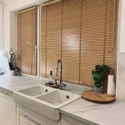 Gama Color - Jasne, drewniane żaluzje poziome w oknie nad zlewem w kuchni. Widoczne detale: marmurowy blat, białe szafki i świeża bazylia w doniczce. Naturalne światło.