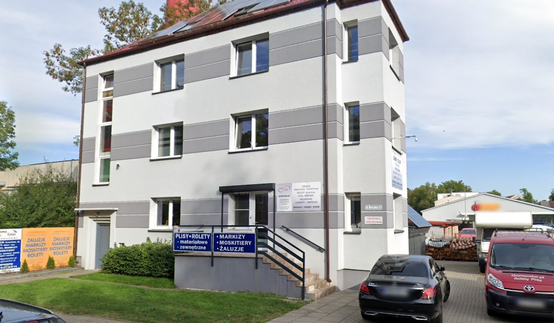 Elewacja budynku z szyldami reklamującymi plisy, rolety, markizy i moskitiery. Widoczne okna i schody wejściowe. Ujęcie z zewnątrz, w dzień.