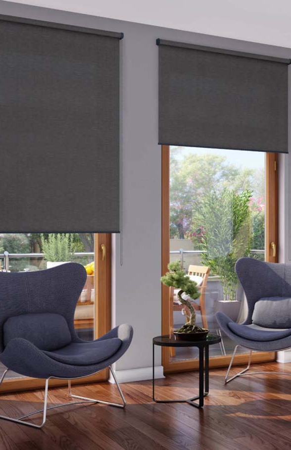 Dwa okna z zasłoniętymi, grafitowymi roletami w nowoczesnym salonie. Widoczne fotele i stolik z bonsai. Naturalne światło z zewnątrz.