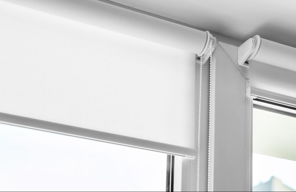 Biała, zwinięta roleta wewnętrzna z łańcuszkiem, zamontowana w oknie. Widoczne mocowania i mechanizm rolety. Delikatna faktura materiału rolety.