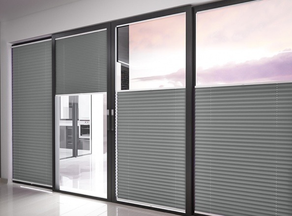 Szare plisy okienne zamontowane w nowoczesnym oknie przesuwnym, widok na kuchnię. Część plis zwinięta, odsłaniając widok na zewnątrz z różowym niebem.