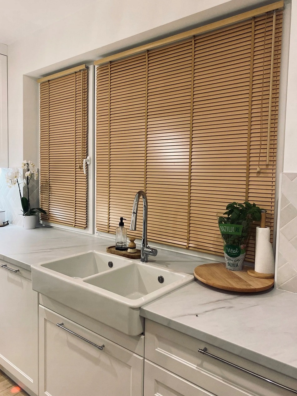 Jasne, drewniane żaluzje poziome w oknie nad zlewem w kuchni. Widoczne detale: marmurowy blat, białe szafki i świeża bazylia w doniczce. Naturalne światło.
