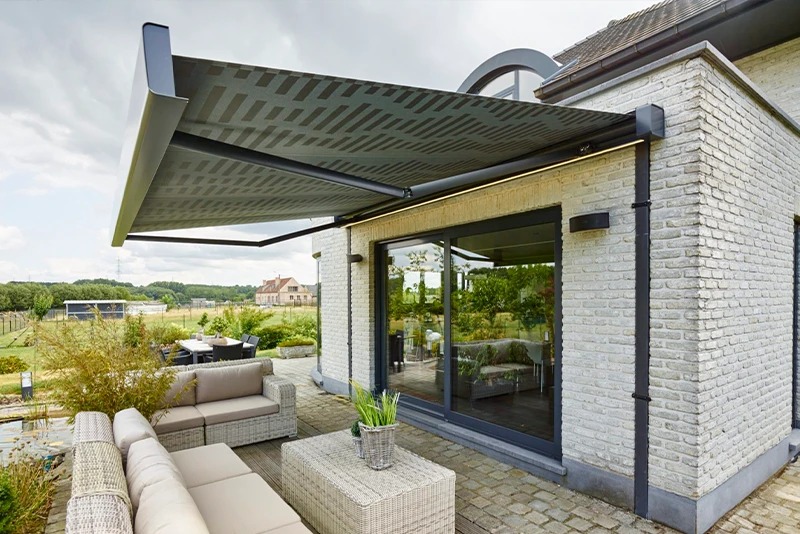 Elegancka markiza tarasowa z szarym wzorem, zamontowana nad przeszklonymi drzwiami, oświetlona listwą LED. W tle ogród i budynek z cegły.