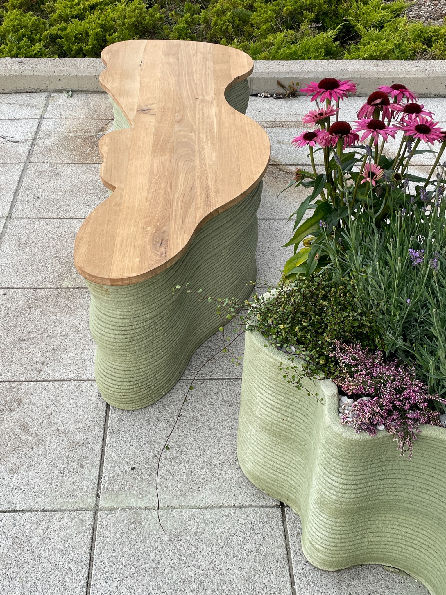 Unikatowa ławka parkowa i donica z kwiatami, wykonane w technologii druku 3D. Ławka z drewnianym siedziskiem i zieloną, warstwową podstawą. Donica w tym samym stylu.