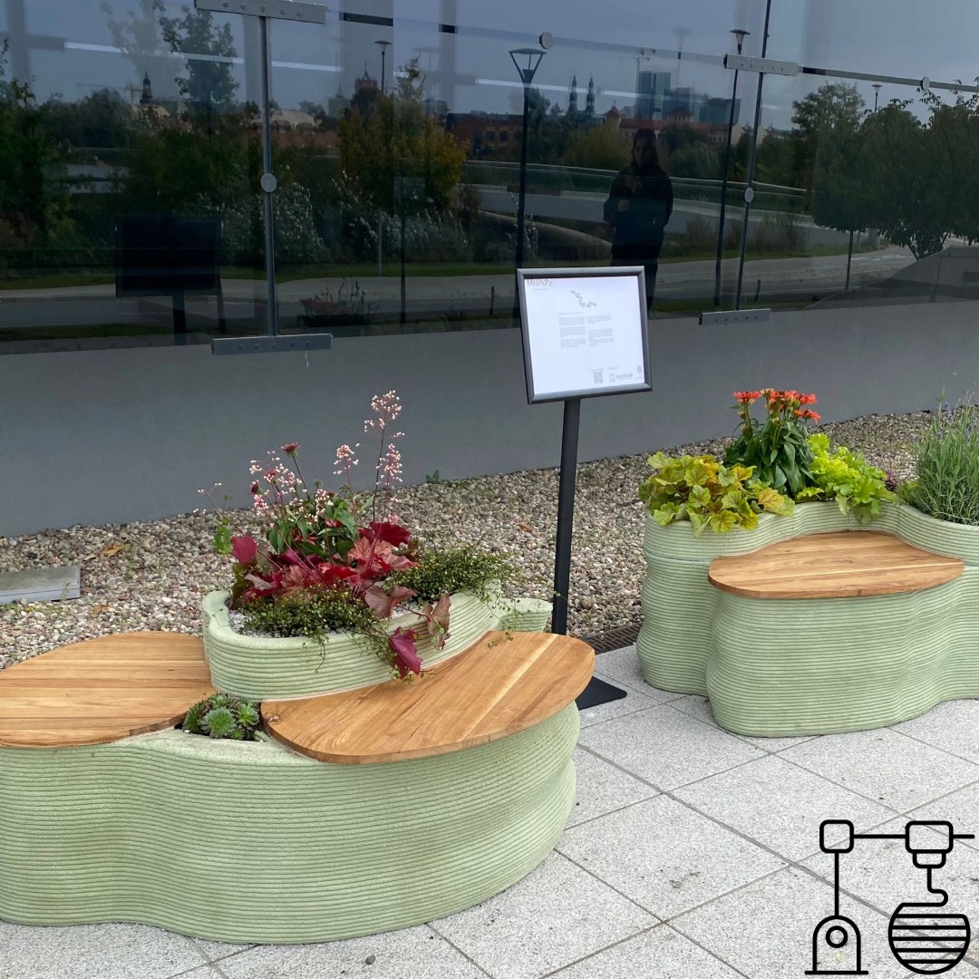 Oryginalne ławki z donicami, wykonane z materiału przypominającego druk 3D, z drewnianymi siedziskami i różnorodną roślinnością, ustawione na zewnątrz budynku.