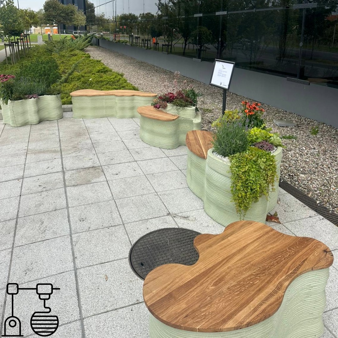 Zestaw ławko-donic z drewna i jasnego tworzywa, uformowanych warstwowo, ustawionych na zewnątrz budynku. Donice obsadzone zielenią i kwiatami.