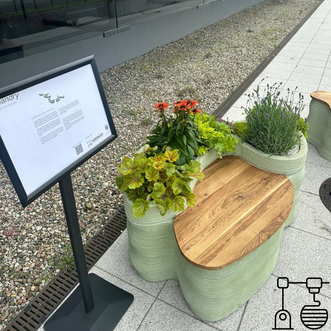 Ławka z donicami, wydrukowana w 3D z jasnego tworzywa, z drewnianym siedziskiem i roślinami. Obok tablica informacyjna z QR kodem. Widok z góry, na zewnątrz.