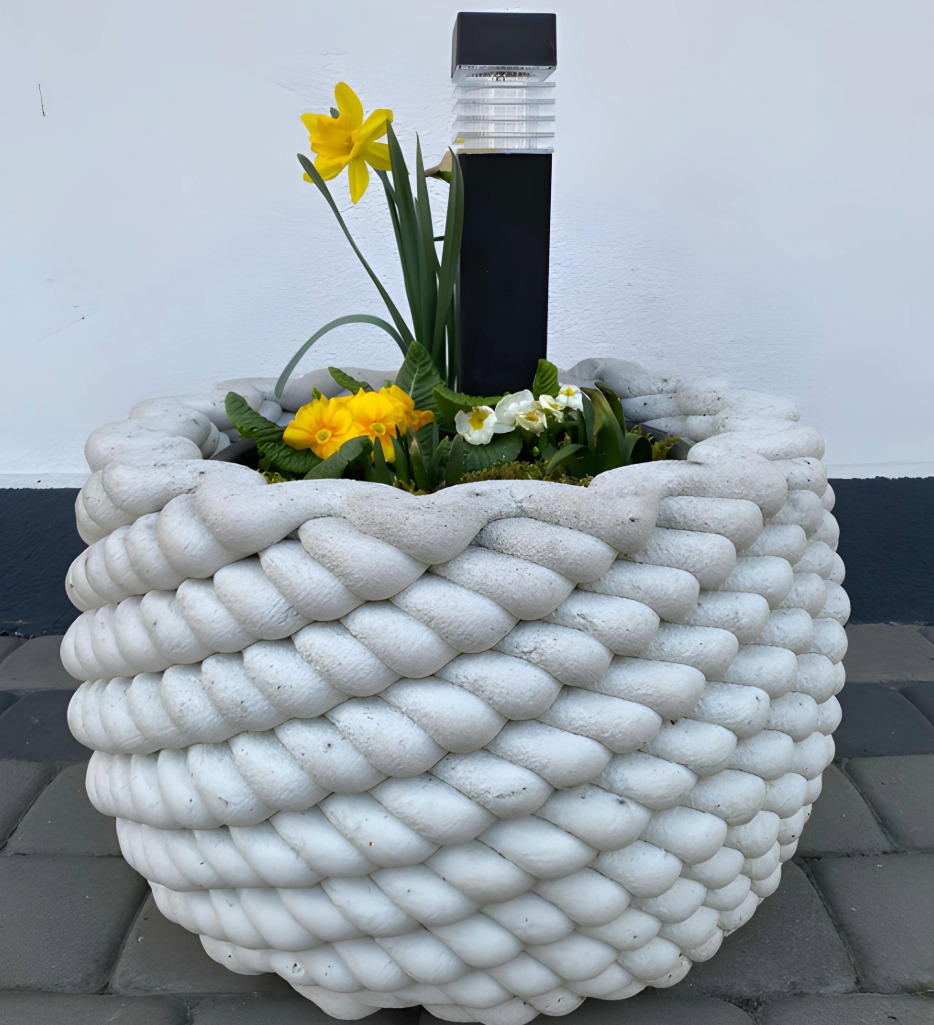 Biała donica z motywem liny, wypełniona kwitnącymi narcyzami i prymulkami, z czarną lampą solarną. Stoi na szarej kostce brukowej, tło stanowi biała ściana.
