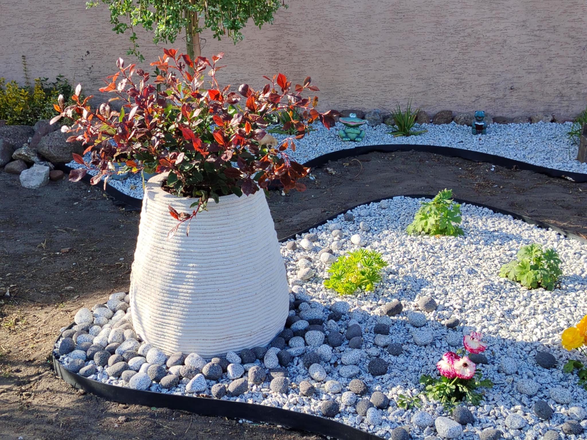Ogród z białym, plecionym wazonem z czerwonolistną rośliną, otoczony szarymi kamieniami i żwirem. W tle trawnik z dekoracjami ogrodowymi.