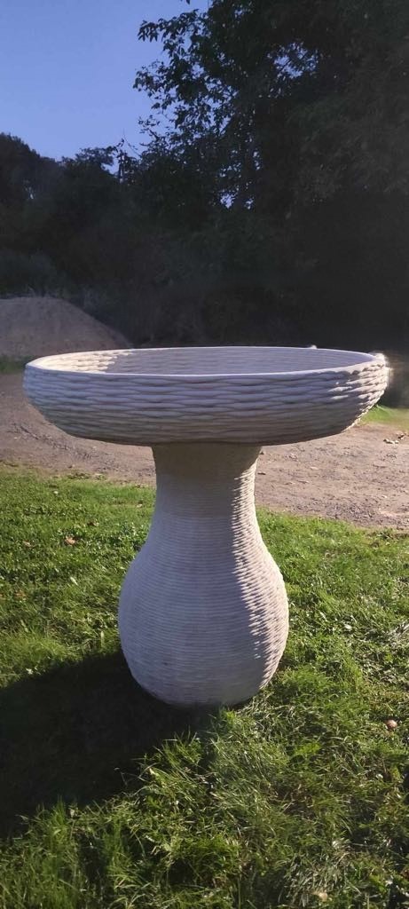 Ozdobny, betonowy stół ogrodowy z wyraźnymi warstwami, prawdopodobnie wykonany metodą addytywną, stoi na trawie na tle drzew i pagórka.