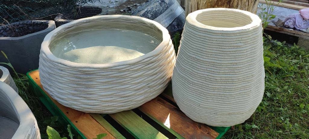 Dwa beżowe, teksturowane wazony/donice, wykonane warstwowo, stoją na drewnianej palecie na zewnątrz. Jeden wazon okrągły, drugi wysoki, w kształcie wazonu.