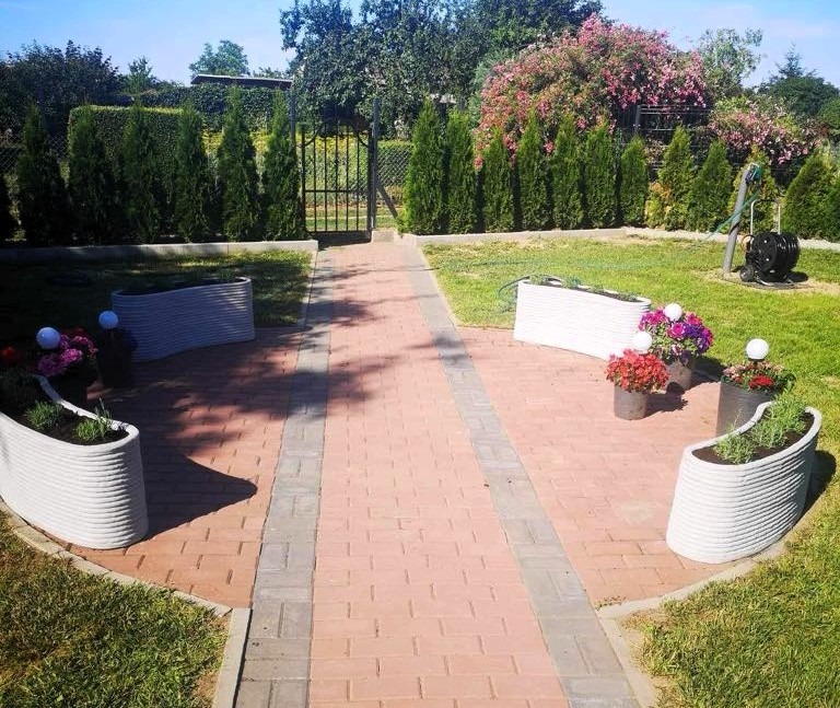 Ogród z ścieżką z kostki i kwietnikami drukowanymi 3D. Widok na bramę i zadbane tuje. Słoneczny dzień, bujna roślinność.