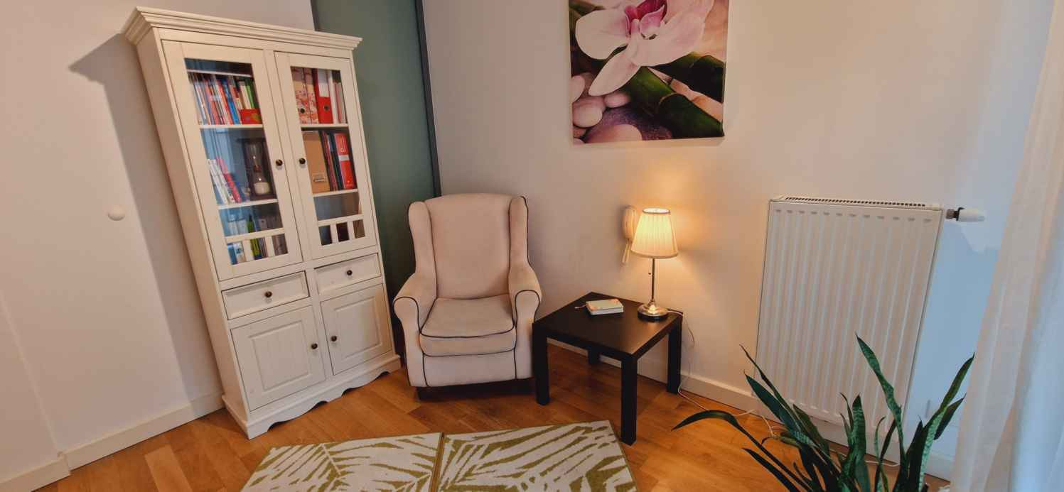 Jasny gabinet: kremowy fotel uszak, regał z książkami, lampka na stoliku, obraz z kwiatem orchidei. Przytulne wnętrze, dywan w liście, drewniana podłoga.