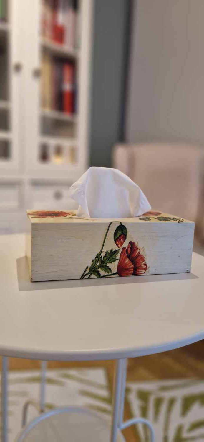 Pudełko na chusteczki z motywem czerwonego maku na białym stoliku w gabinecie. W tle regał z książkami i fotel. Przytulne wnętrze poradni.