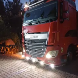 Czerwona ciężarówka marki DAF z włączonymi światłami, zaparkowana na brukowanej nawierzchni o zmierzchu. Widoczne drzewa w tle.