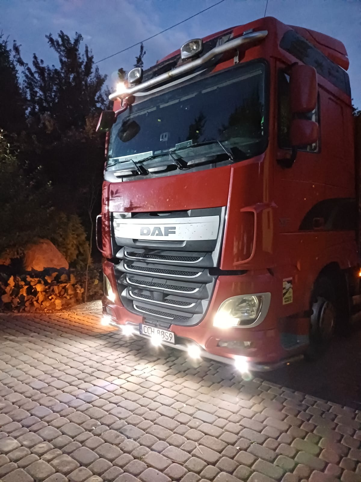 Czerwona ciężarówka marki DAF z włączonymi światłami, zaparkowana na brukowanej nawierzchni o zmierzchu. Widoczne drzewa w tle.