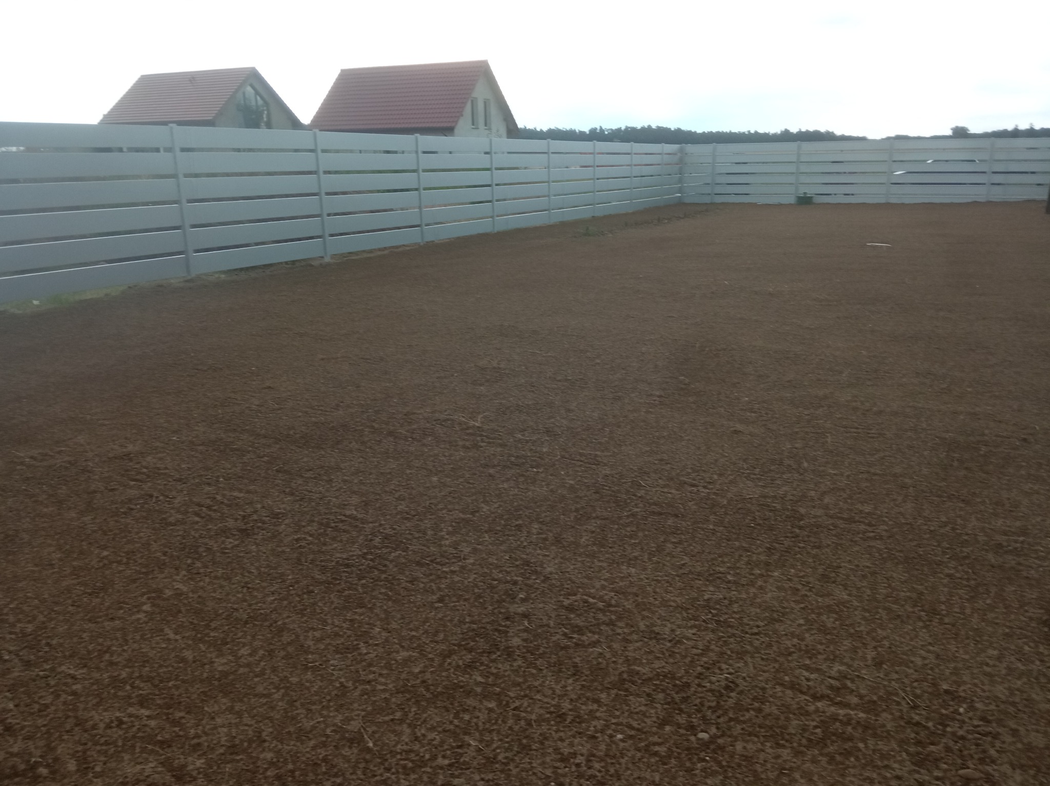 Przygotowany teren pod trawnik, wyrównana ziemia, otoczony białym, nowoczesnym ogrodzeniem panelowym. W tle widoczne budynki mieszkalne.