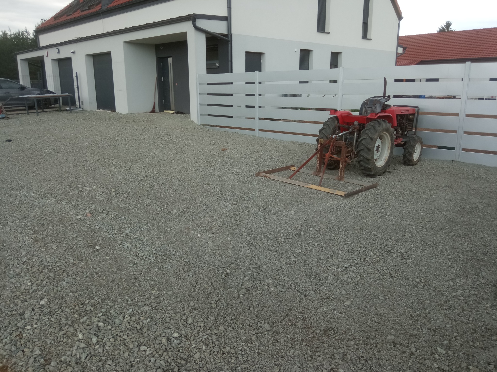 Czerwony traktor z broną na żwirowym podłożu przed nowoczesnym domem z szarym ogrodzeniem. Przygotowanie terenu pod ogród.