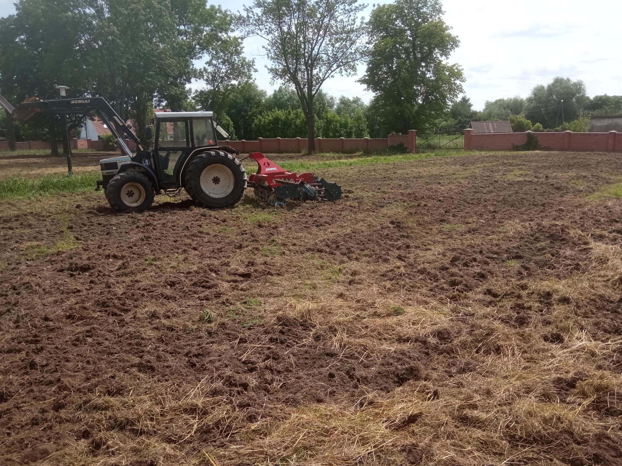 Ciągnik z glebogryzarką na polu, przygotowane podłoże, widoczny mur z czerwonej cegły w tle. Prace ziemne.