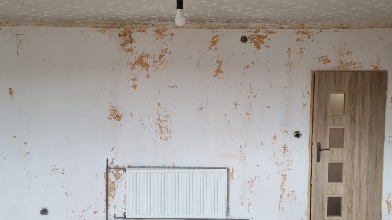 Pokój przed remontem: ściany z resztkami farby, grzejnik, drzwi z szybkami. Widoczne ślady po szpachlowaniu i przygotowaniu do malowania. Żarówka zwisa z sufitu.