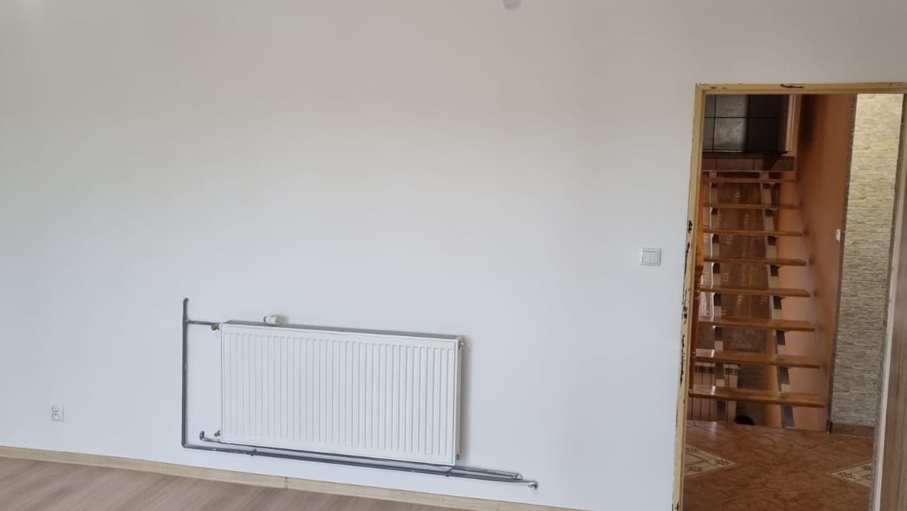 Biała ściana po malowaniu, widoczny grzejnik i schody w głębi. Minimalistyczny wystrój wnętrza po odświeżeniu. Jasne, neutralne barwy.