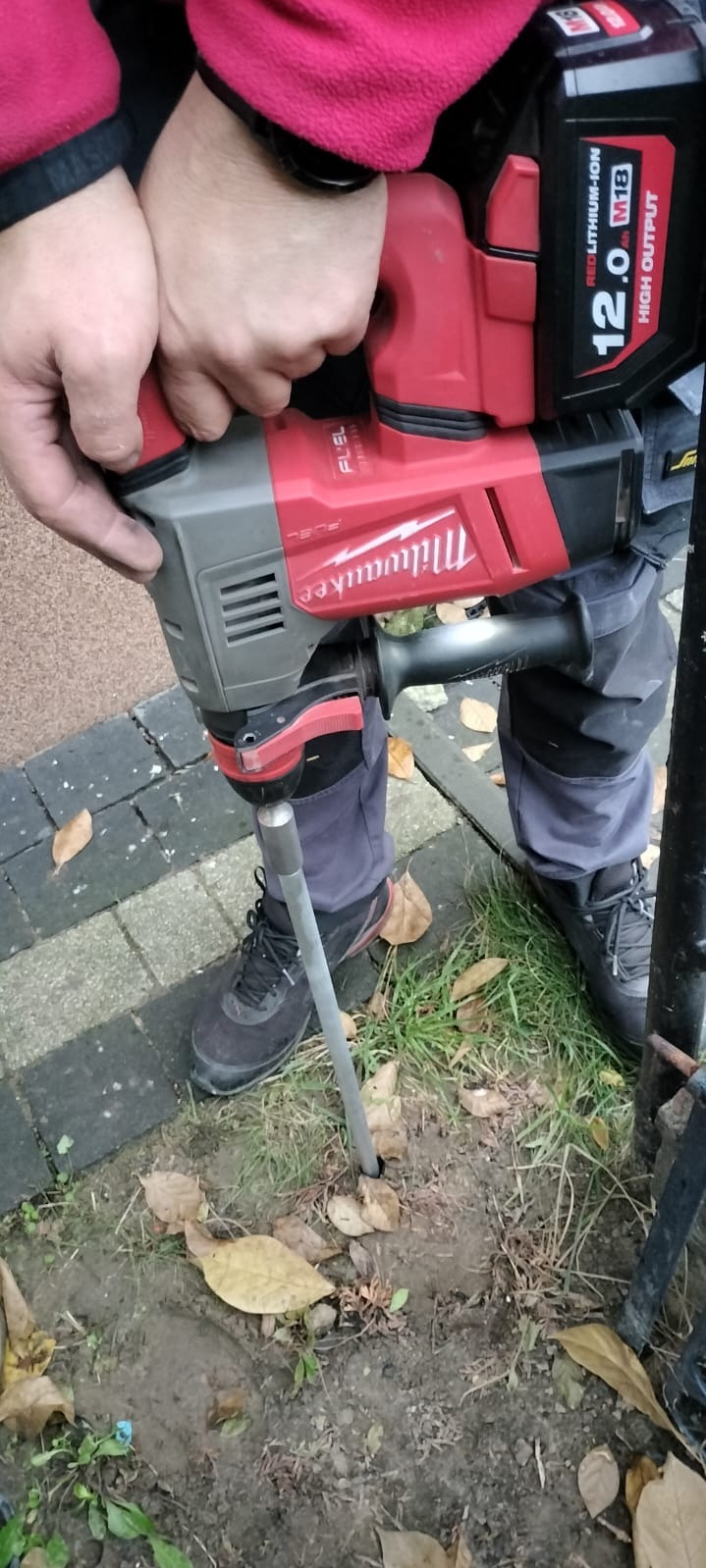 Wbijanie szpilki uziemiającej młotem udarowym Milwaukee. Widoczne ręce operatora, narzędzie i fragment wbitej szpilki w ziemi. Prace ziemne przy ogrodzeniu.