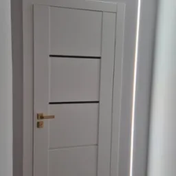 Nowoczesne wykończenie wnętrza: białe drzwi z złotą klamką i dekoracyjne oświetlenie LED w kształcie litery L na szarej ścianie, czarny reflektor sufitowy.