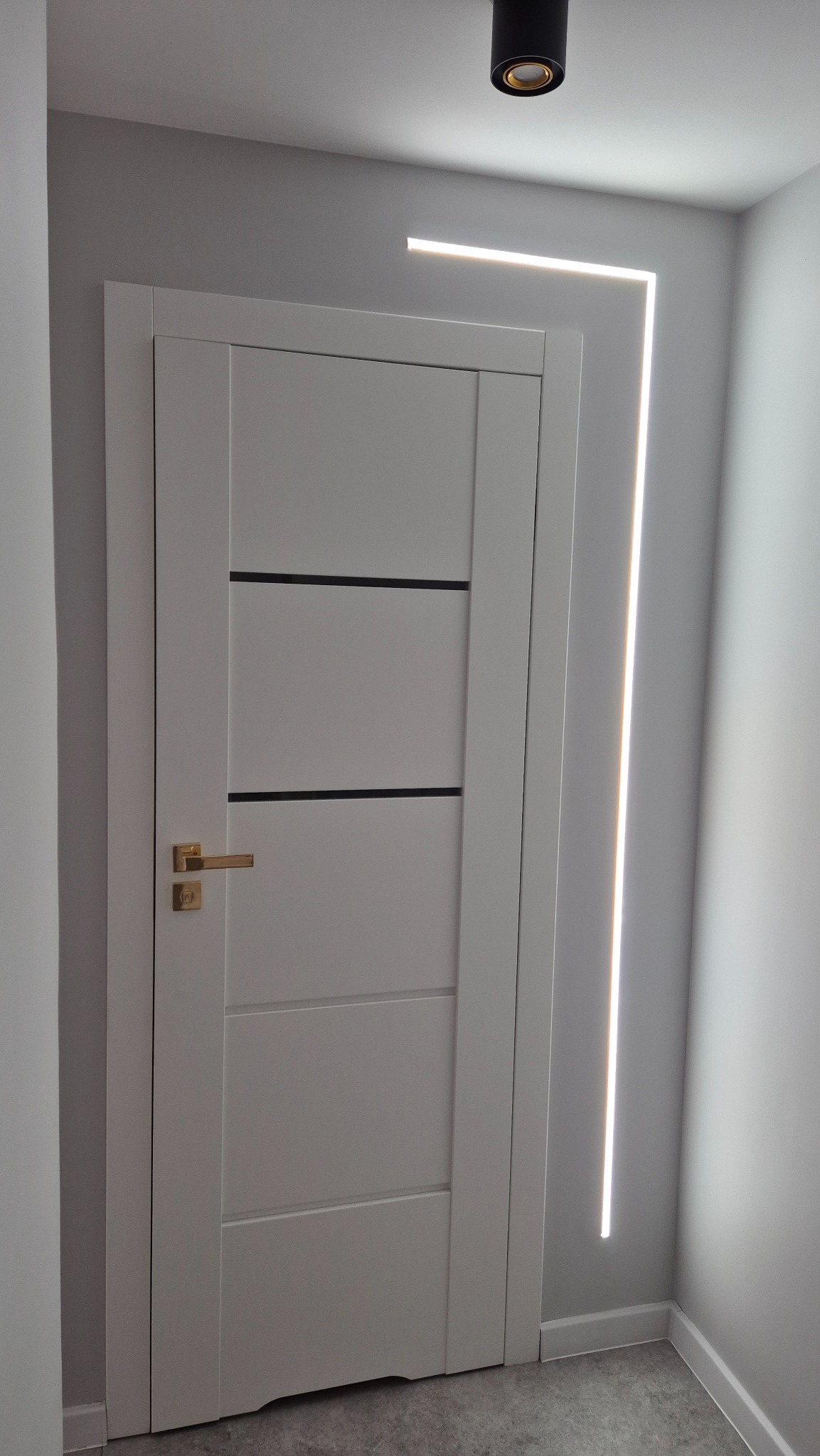 Nowoczesne wykończenie wnętrza: białe drzwi z złotą klamką i dekoracyjne oświetlenie LED w kształcie litery L na szarej ścianie, czarny reflektor sufitowy.