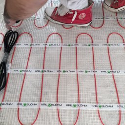 Kril & Blöhm - Montaż maty grzewczej w Warszawie. Czerwone kable układane na białej siatce, widoczne stopy w trampkach. Instalacja ogrzewania podłogowego w trakcie realizacji.