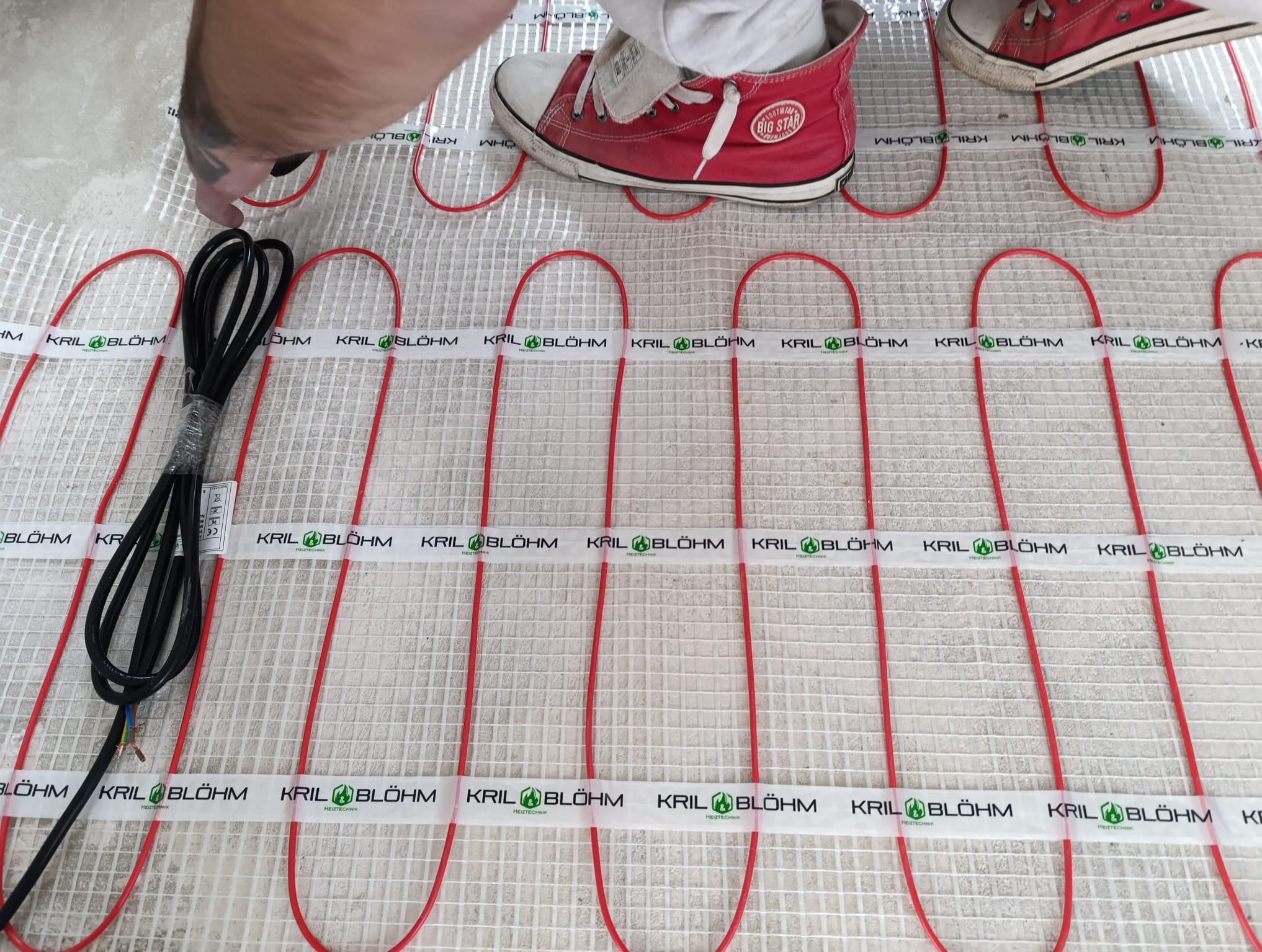 Montaż maty grzewczej w Warszawie. Czerwone kable układane na białej siatce, widoczne stopy w trampkach. Instalacja ogrzewania podłogowego w trakcie realizacji.