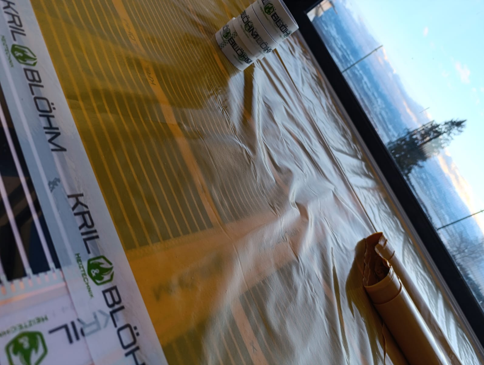 Montaż maty grzewczej Kril&Blöhm na podłodze, widok z góry, z rolką kabla i fragmentem okna z widokiem na zewnątrz. Delikatne światło dzienne.