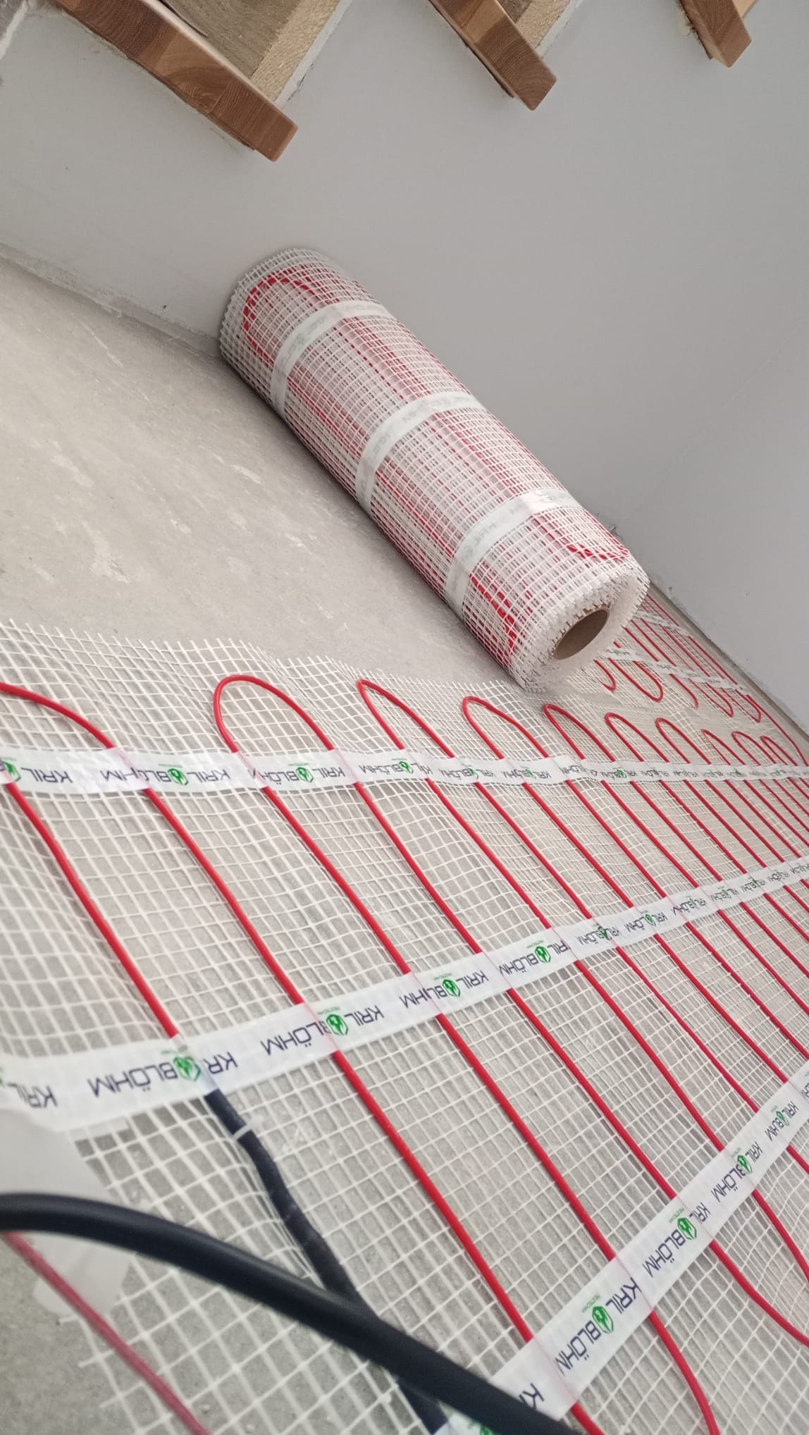 Rozwinięta mata grzewcza z czerwonymi przewodami na siatce, częściowo w rolce. Widoczne przewody zasilające. Montaż ogrzewania podłogowego.