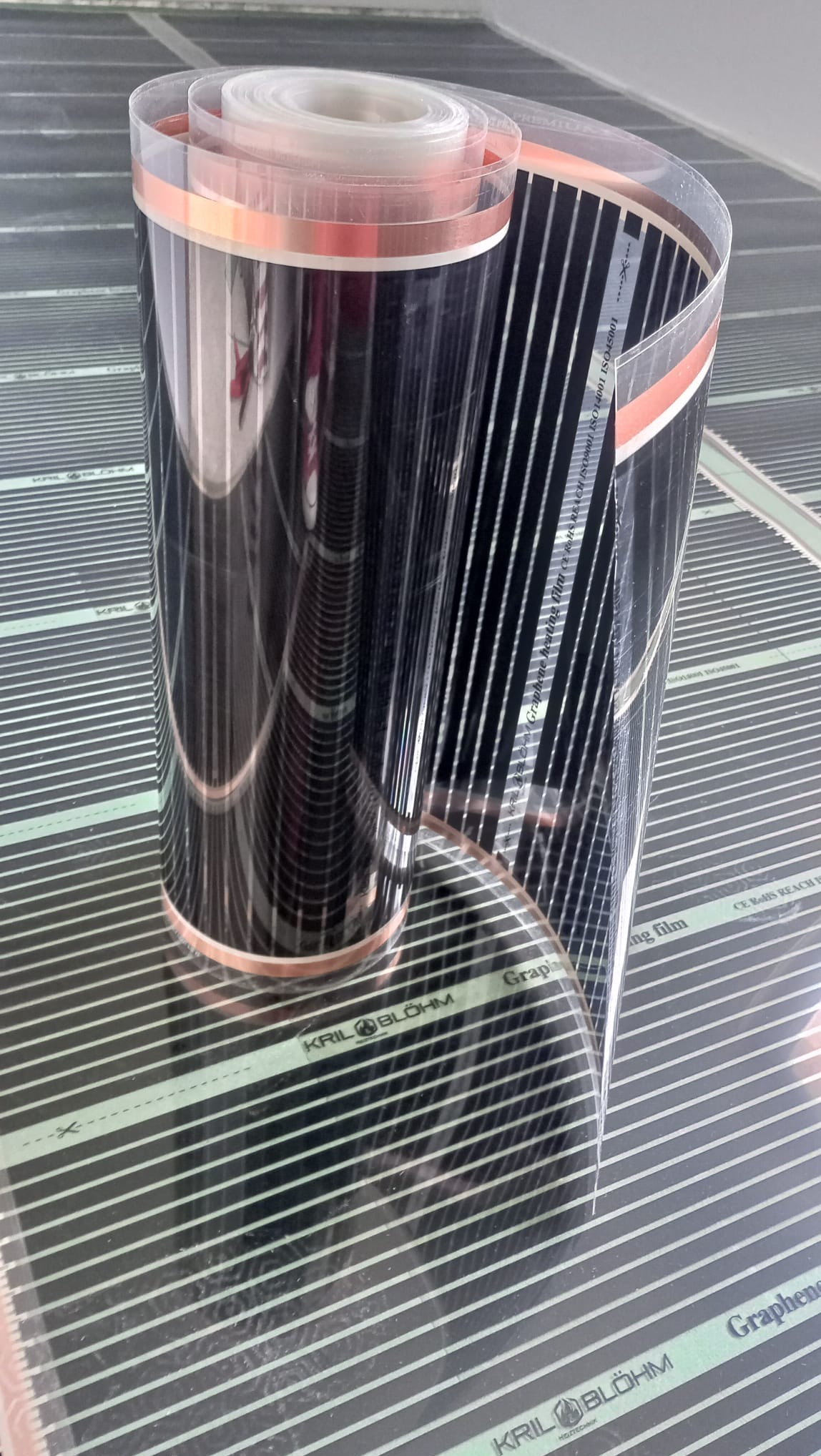 Rolka czarnej maty grzewczej z widocznymi liniami grzewczymi, leżąca na rozłożonej macie tego samego typu. Widoczne oznaczenia producenta.