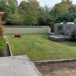 Zadbany trawnik z obrzeżem z kostki, fontanną z kamienia i altaną w tle. Widoczne rośliny w donicach i mała walcarka do trawy. Całość tworzy estetyczny ogród.