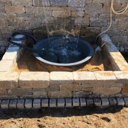 LUCABAU GARTENBAUPROFI WIESŁAW WALCZAK - Fontanna z kamienia naturalnego w ogrodzie, otoczona kostką granitową. Elementy instalacji wodnej widoczne. Całość w surowym, naturalnym stylu.